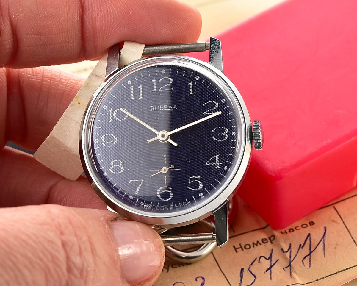 Pobeda-10448_KOM_1949 Hand holding a vintage mechanical wristwatch with blue dial, shown next to red box and paperwork.